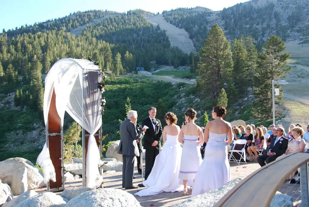 A wedding ceremony unfolds outdoors, framed by mountains and pine trees. The bride and groom stand before an officiant under a decorated arch, while guests in formal attire are seated on white chairs. Bathed in natural sunlight, this picturesque scene captures the essence of a perfect wedding day. Mt. Rose Ski Tahoe