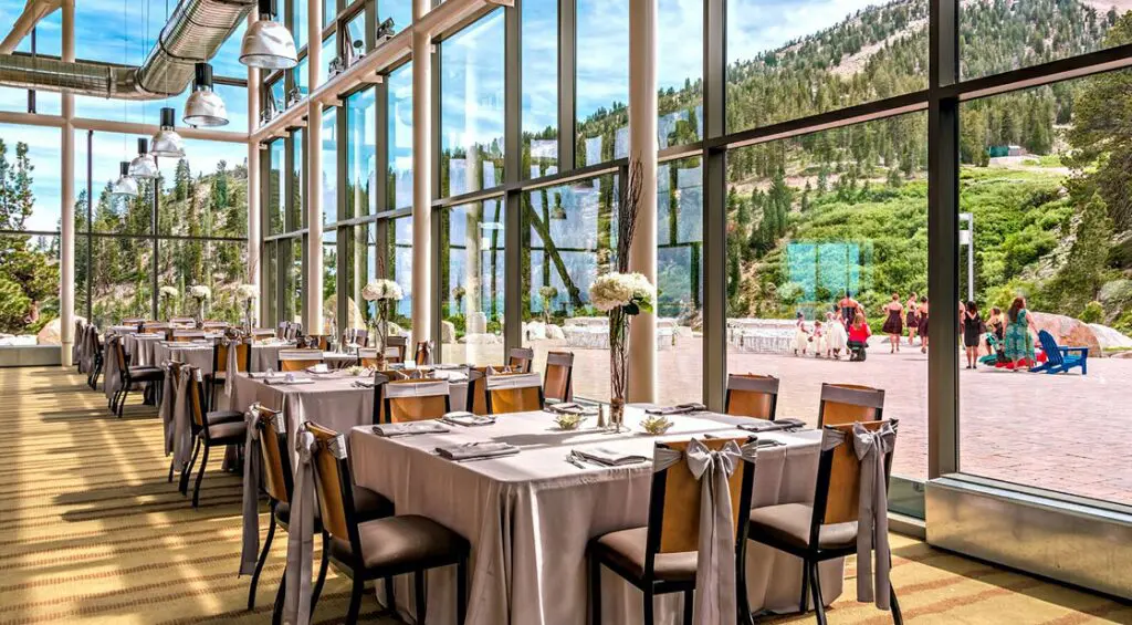 A bright, spacious room with large floor-to-ceiling windows overlooking a mountainous landscape. Multiple tables are elegantly set with white tablecloths and floral centerpieces. Outside, a wedding ceremony is taking place among lush greenery and scenic views. Mt. Rose Ski Tahoe