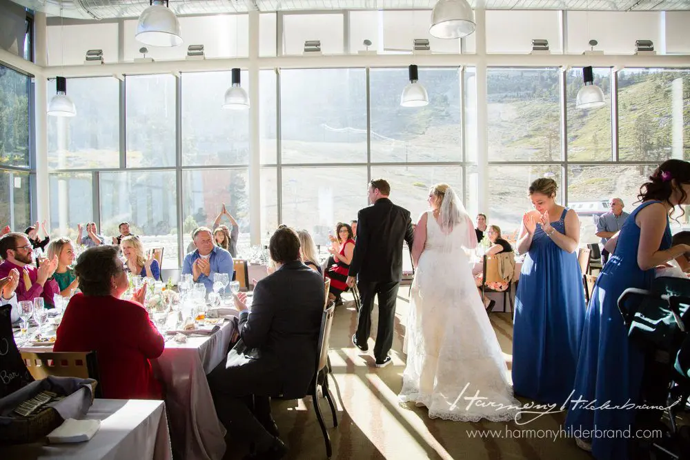 A bride and groom walk hand in hand through a sunlit wedding reception inside a spacious venue with large windows. Guests, seated at decorated tables, applaud the couple. The bridal party, dressed in blue, stands nearby, basking in the bright, natural light. Mt. Rose Ski Tahoe