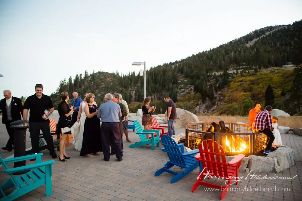 A group of people socialize around a fire pit outdoors in a mountainous area. They are gathered on a patio with colorful Adirondack chairs. The scene is set during evening time, and greenery-covered hills are visible in the background. Mt. Rose Ski Tahoe