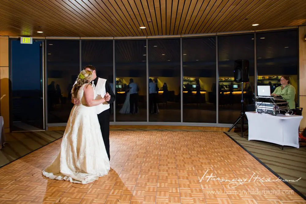A bride and groom share their first dance in a dimly lit reception hall with wooden paneling. They are centered on a parquet dance floor, with guests and a DJ in the background near large windows reflecting the interior lights. Mt. Rose Ski Tahoe