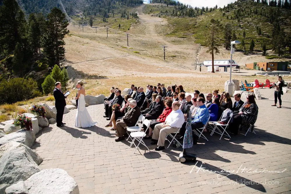 A wedding ceremony takes place outdoors in a mountainous area. The bride and groom stand at the altar, with guests seated on white folding chairs. The background features ski lifts, dry grass, and a few scattered trees under a clear sky. Mt. Rose Ski Tahoe