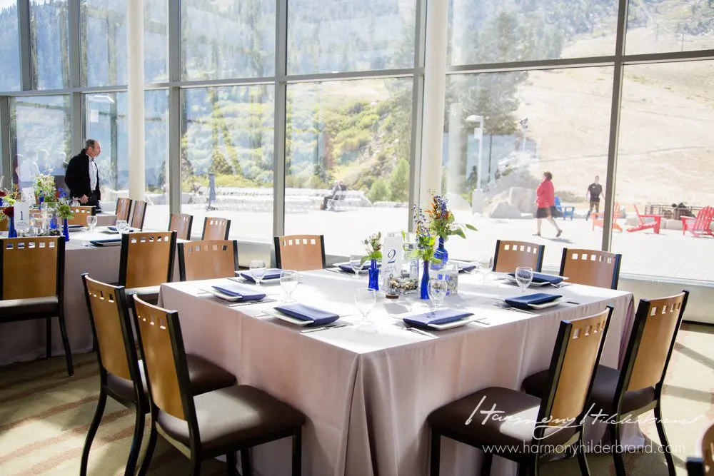 A beautifully set dining table is in the foreground with white tablecloths, navy blue napkins, and elegant tableware. Large windows behind showcase a scenic mountainous landscape with greenery. Two people are visible in the background, next to additional seating. Mt. Rose Ski Tahoe