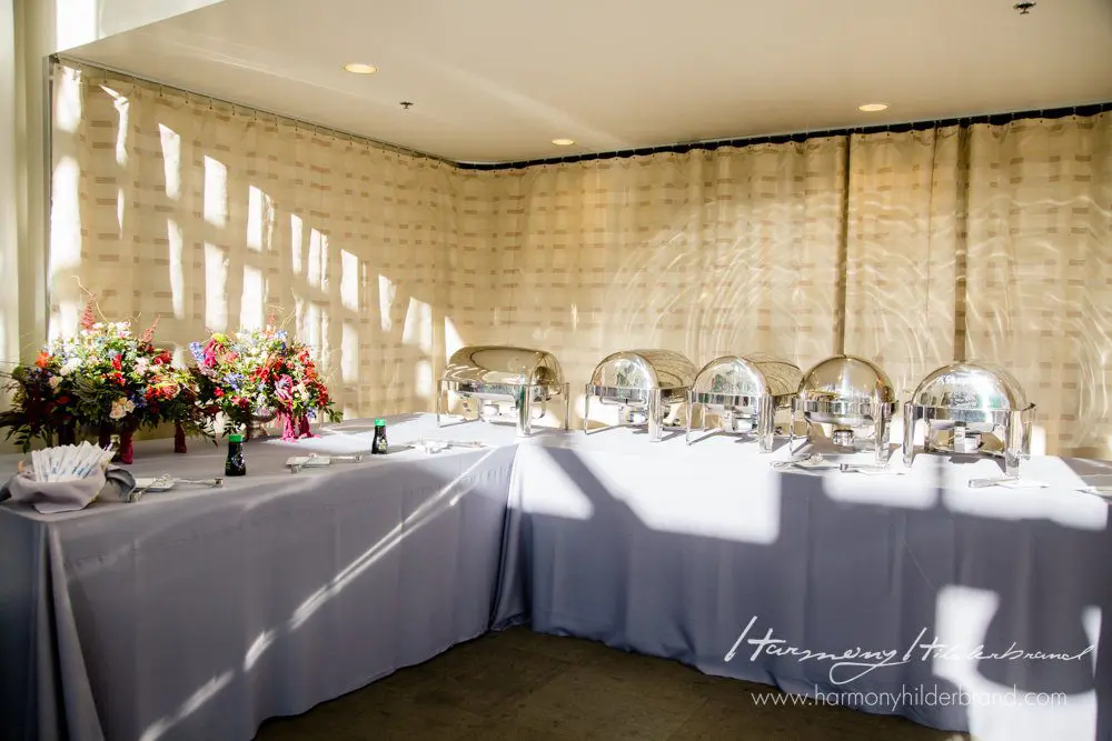 A buffet setup in a bright room with a long table covered in white tablecloths. Five chafing dishes with lids are arranged in a row, accompanied by a floral arrangement, bottles, and folded napkins. The background features a cream-colored curtain with sunlight filtering through. Mt. Rose Ski Tahoe