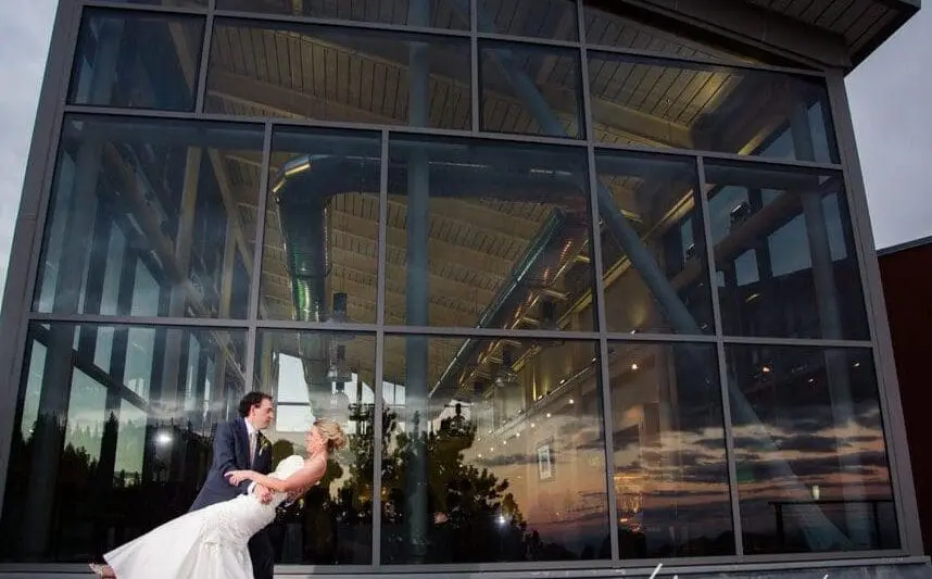 A couple in wedding attire is dancing outside a modern building with large reflective windows. The groom dips the bride, who holds a bouquet, as they smile. The setting sun and interior lights of the building add a warm glow to the scene. Mt. Rose Ski Tahoe