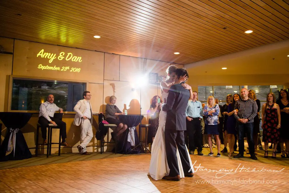 A bride and groom share their first dance at their wedding reception. Guests stand around the dance floor, watching and smiling. The wedding date, "September 23rd, 2016," and names "Amy & Dan" are projected on the wall. The venue has a warm, lit ambiance. Mt. Rose Ski Tahoe