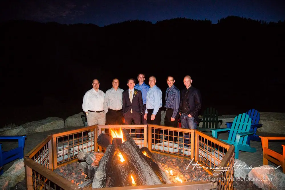 A group of seven people stands around a campfire at night. The fire is enclosed by a metal mesh barrier, and there are colorful Adirondack chairs around the area. Behind them, trees and hills are silhouetted against the dark sky. Mt. Rose Ski Tahoe