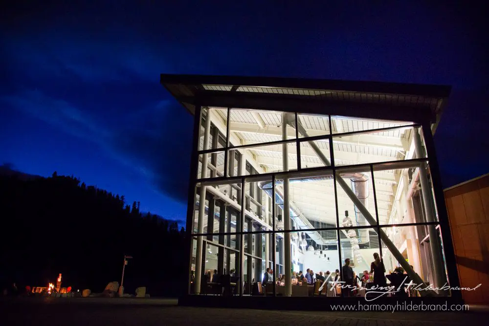 A modern, glass-walled building, illuminated from within, stands against a deep blue evening sky. Through the windows, people can be seen engaging in activities inside. The scene exudes a warm and inviting atmosphere. The website harmonyhildebrand.com appears in the bottom right corner. Mt. Rose Ski Tahoe