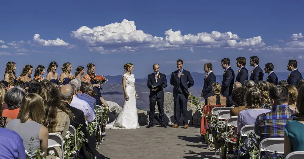 Outdoor wedding ceremony on a clear sunny day with a scenic mountain backdrop. The bride and groom stand in the center, flanked by bridesmaids in orange dresses on the left and groomsmen in dark suits on the right. Guests are seated, facing the couple. Mt. Rose Ski Tahoe