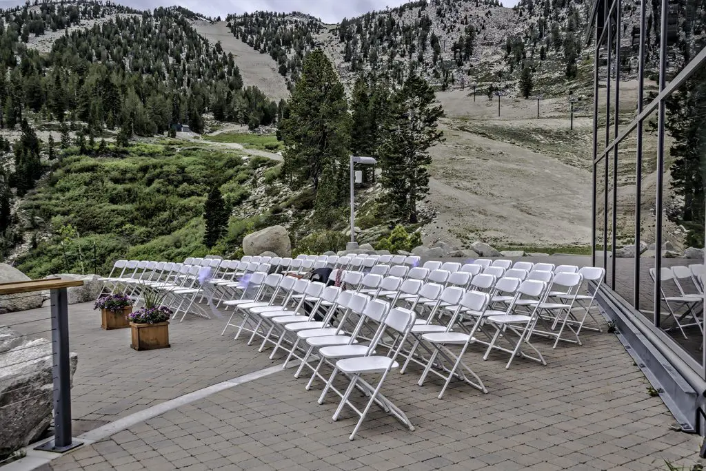 Rows of white folding chairs are set up in an outdoor ceremony space on a paved area. The backdrop features a mountainside covered in greenery and pine trees. The chairs face a reflective glass building, and flower arrangements in wooden planters are placed along the aisle. Mt. Rose Ski Tahoe