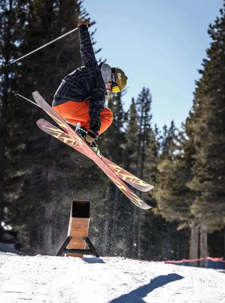 Mt. Rose A skier in mid-air performs a trick over a small ramp in a snowy forested area. The skier is wearing an orange and black outfit with a helmet and goggles. Sunlight filters through the trees in the background.