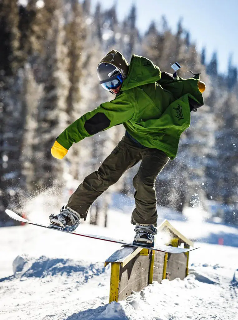 Mt. Rose A snowboarder wearing a green jacket and helmet performs a trick on a rail in a snowy landscape with tall, snow-covered trees in the background. The snowboarder is balancing with one hand stretched out and snow spraying off the rail.