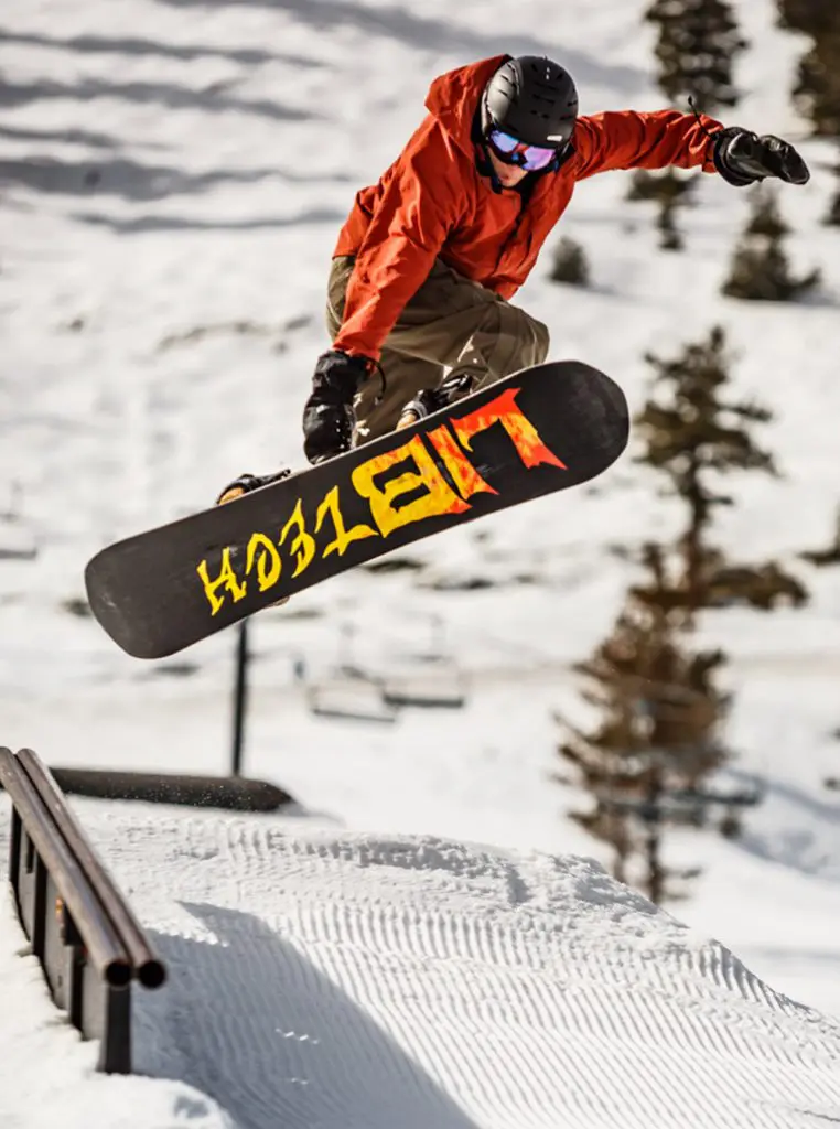 Mt. Rose A snowboarder in an orange jacket and black helmet performs a trick mid-air over a snow-covered slope. The snowboard, displaying bold yellow and orange graphics, is prominently seen as the person rides slightly above a rail. Snowy mountains and trees are in the background.