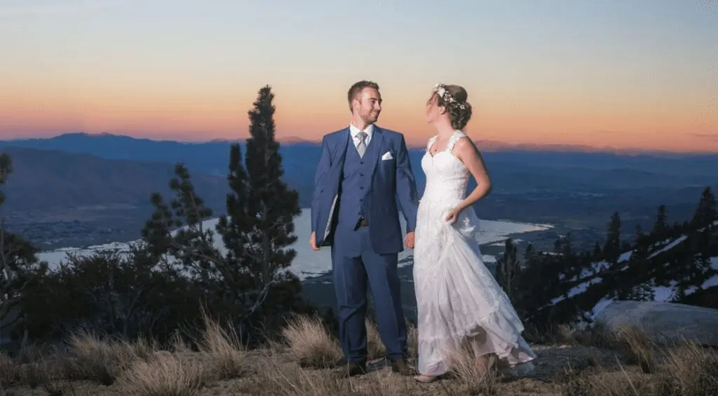 A bride and groom stand hand in hand on a rocky hillside at sunset, enveloped in the magic of their wedding day. The groom wears a blue suit, while the bride is in a white lace gown with a floral headpiece. Behind them, a panoramic view of valleys, trees, and a distant water body under a colorful sky is visible. Mt. Rose Ski Tahoe