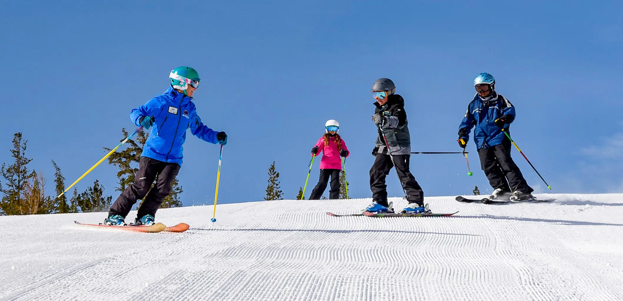 A group of four people, including children and adults, are skiing down a snowy slope on a sunny day. They are all wearing winter gear, including helmets, jackets, and ski boots. There are colorful markers on the snow from the best kids ski lessons, tall pine trees, and a clear blue sky in the background. Mt. Rose Ski Tahoe