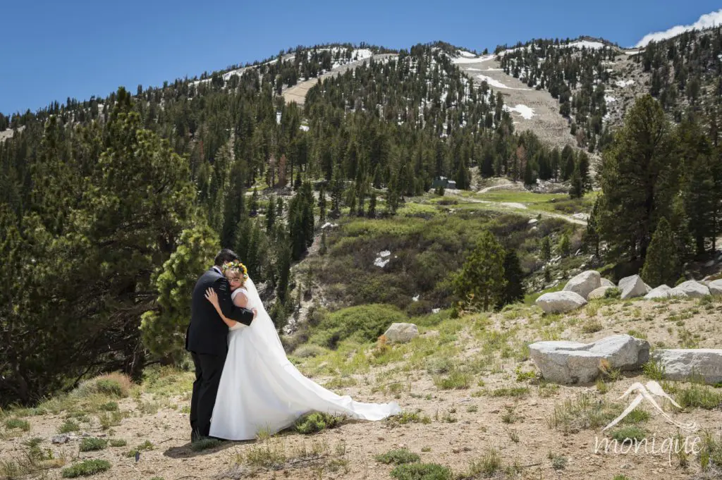 A bride and groom embrace in a mountainous landscape with scattered trees and patches of snow. The bride wears a white dress with a long train and floral headpiece while the groom wears a black suit. The clear blue sky forms the perfect backdrop for their wedding day. "imonie" is written in the bottom right. Mt. Rose Ski Tahoe