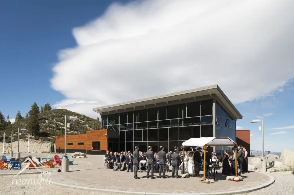 A beautiful wedding ceremony is taking place outdoors in front of a modern glass building surrounded by scenic mountains. Guests are seated facing the couple and officiant under a white canopy. The sky is clear with some clouds, and trees are visible in the background. Mt. Rose Ski Tahoe
