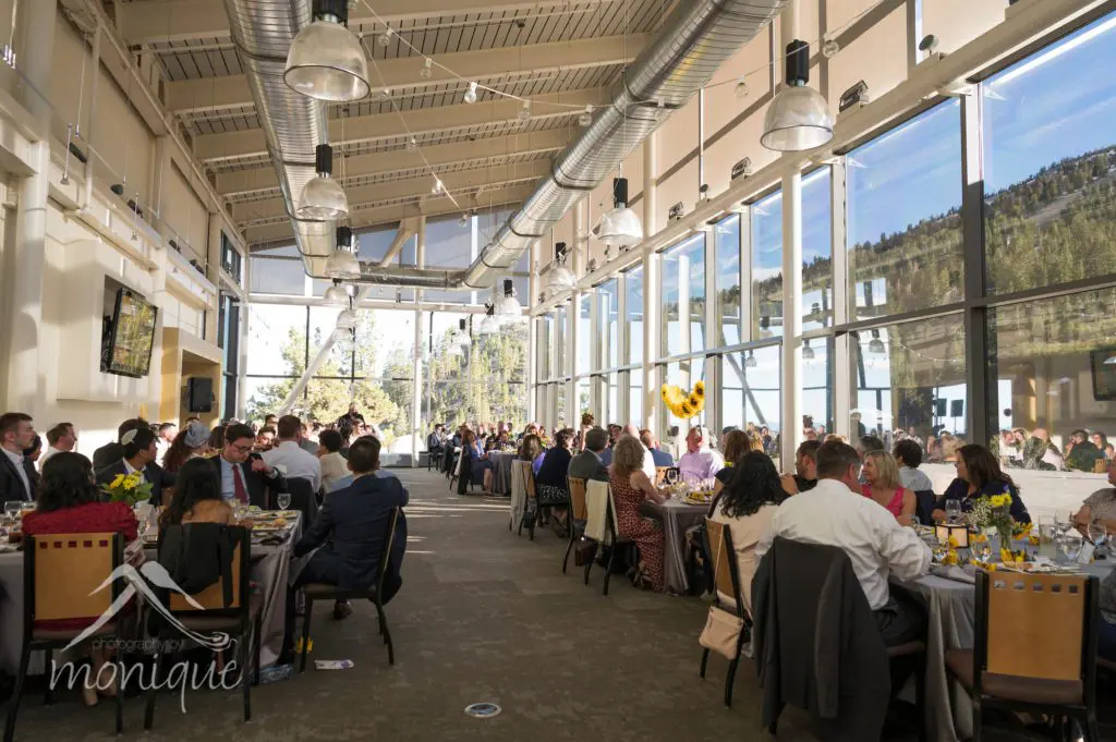 A large wedding gathering is seated at tables in a well-lit room with large windows offering a mountain view. The space is decorated with sunflower centerpieces. The ceiling features industrial-style ductwork, and natural light beautifully fills the room. Mt. Rose Ski Tahoe