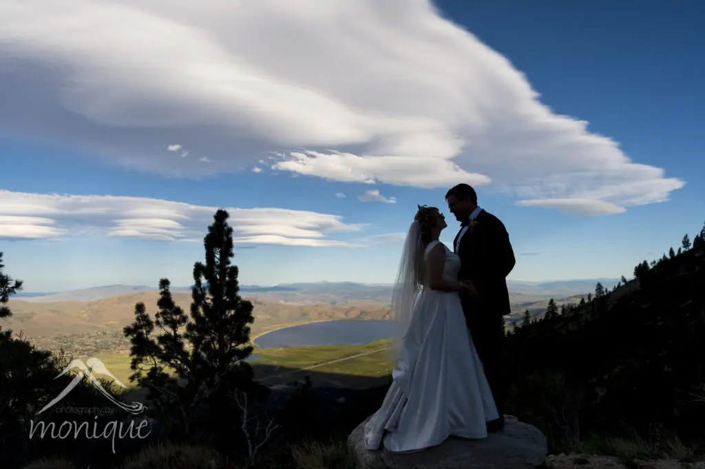 The wedding couple stands on a rock overlooking a scenic landscape with rolling hills, a distant lake, and dramatic cloud formations in the sky. The bride, in her flowing white dress and veil, shares an intimate moment with the groom dressed sharply in his suit. Mt. Rose Ski Tahoe