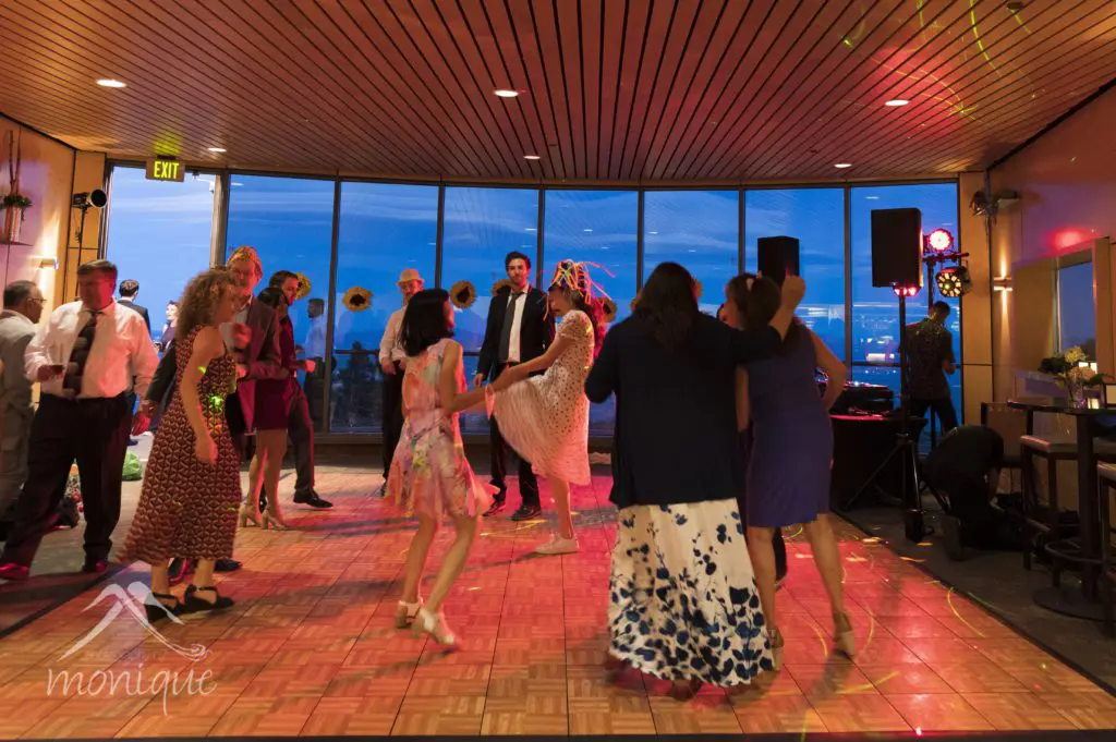 People are dancing in a warmly lit room with large windows and a wood-paneled ceiling, celebrating a wedding. A few disco lights add colorful accents to the setting. The floor is wooden, and there’s a small DJ setup on the right. The background shows an evening cityscape. Mt. Rose Ski Tahoe