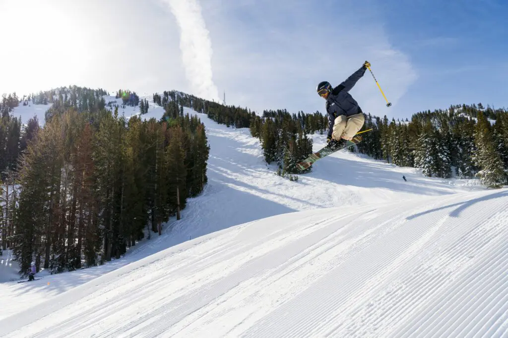 Mt. Rose A skier performs a jump trick on a sunny day at Ski Tahoe. The skier is airborne, holding their ski poles out to the sides, with trees and mountainous slopes in the background. The snow is freshly groomed, adding to the pristine conditions.
