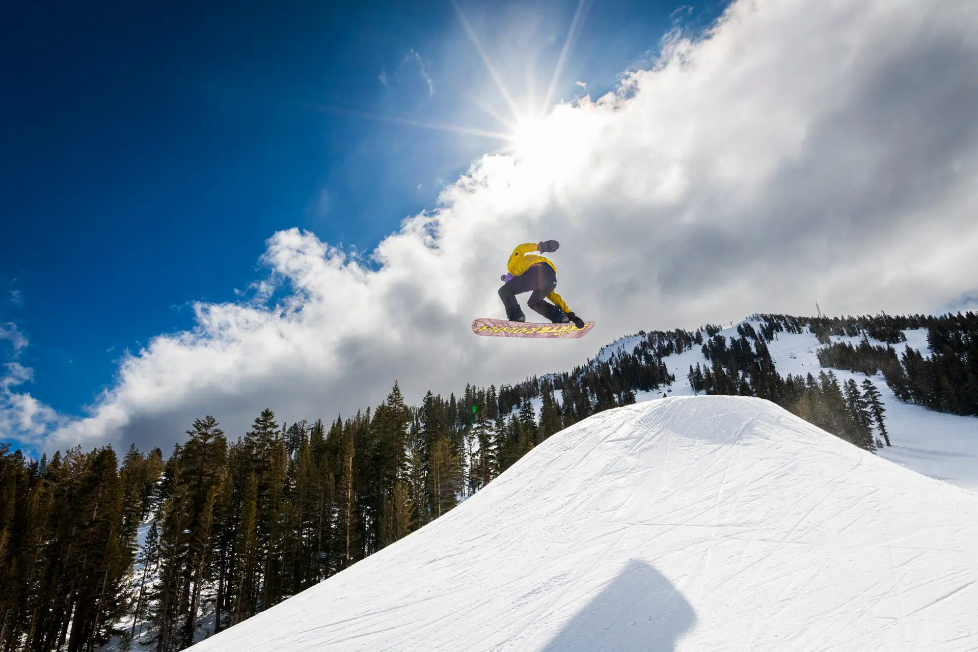 Mt. Rose A snowboarder in a yellow jacket catches air while performing a jump on a snow-covered slope surrounded by pine trees. The bright sun shines through partially cloudy skies, casting a striking light over the winter landscape at Ski Tahoe.