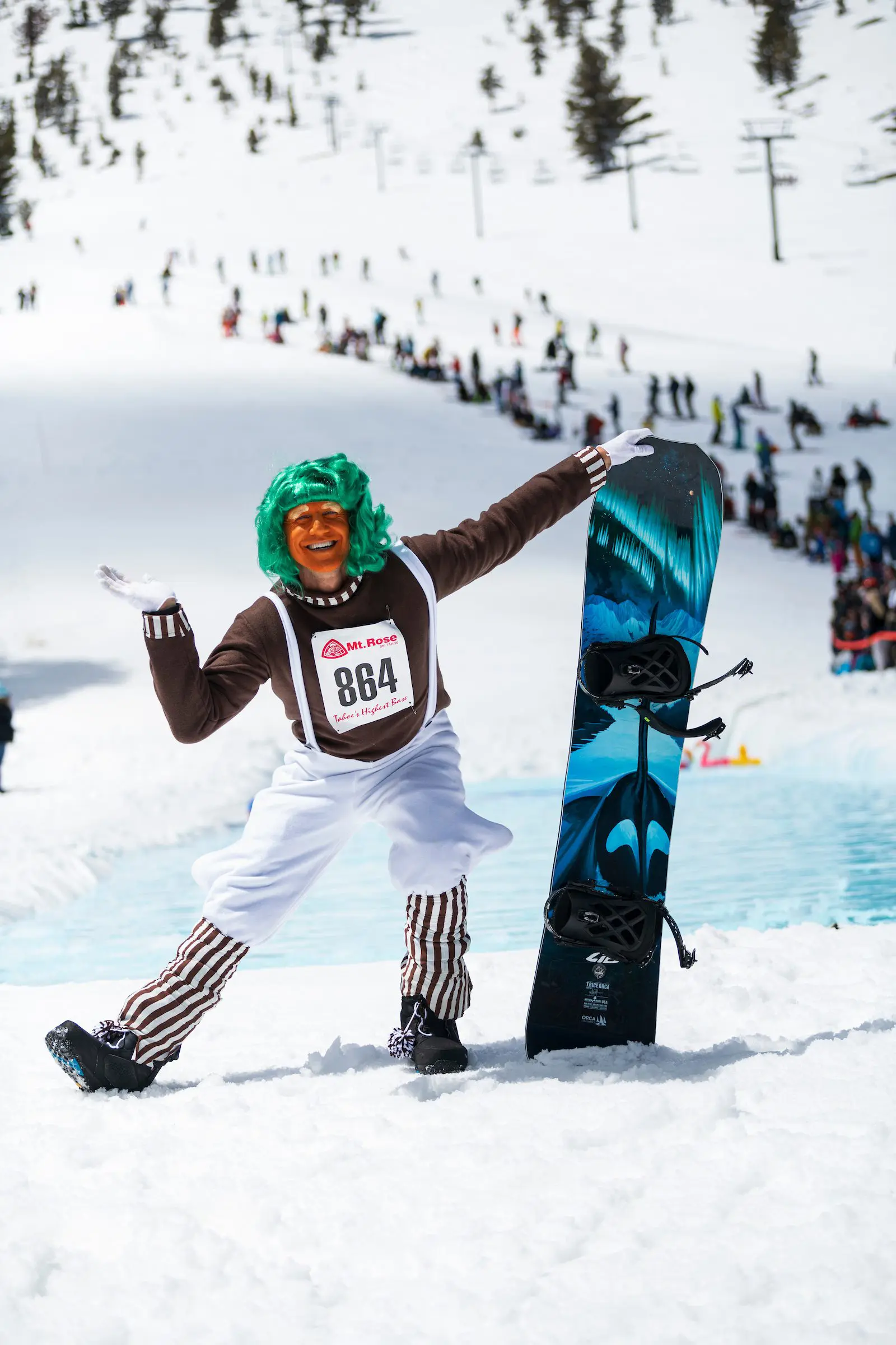 Mt. Rose A person dressed in an Oompa Loompa costume with green hair, brown shirt, white overalls, and striped leggings poses with a snowboard beside a snow-covered slope. They wear a bib labeled "864", and a crowd is visible in the background.