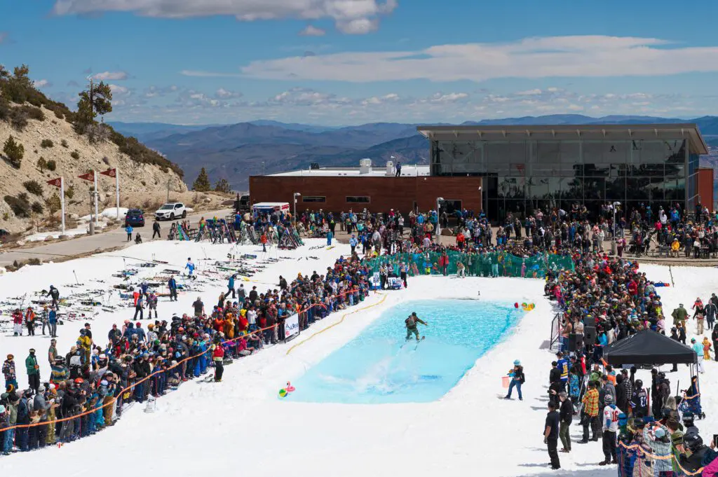 Mt. Rose A busy ski resort with a large crowd gathered around a rectangular pool on the snowy slopes of Slide Mountain. Skiers and snowboarders are in line for chairlifts, and ski equipment is laid out on the snow. Snow-covered trees and mountains form a picturesque backdrop under a blue sky near Winters Creek Lodge.