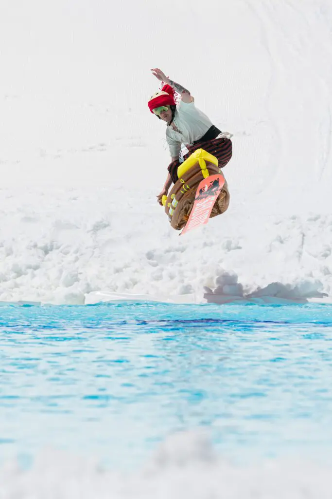 Mt. Rose A person in a red helmet and white shirt is snowboarding on snow but has just jumped over a section of bright blue water. The background is white snow, and the rider is mid-air with a snowboard angled to the side.
