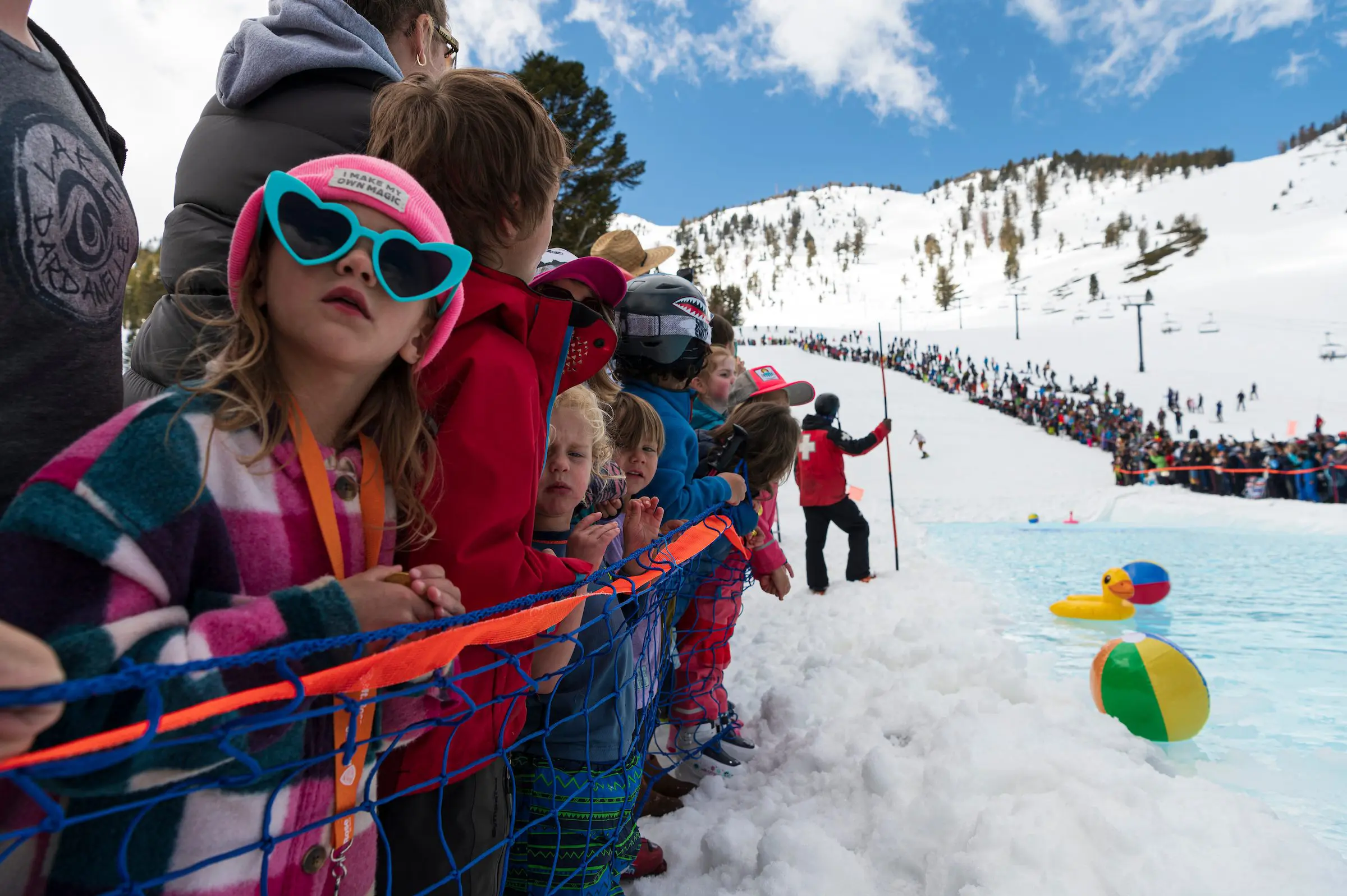 Mt. Rose Children in colorful winter outfits and sunglasses stand behind a safety net, eagerly watching a snowy ski event. The crowd stretches into the snowy landscape, with a pool and floating beach balls in the foreground and ski lifts visible in the background.