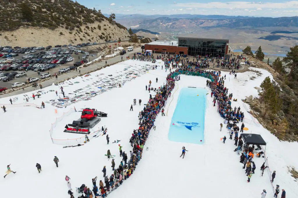 Mt. Rose A large crowd gathers around a rectangular pool set up on a snowy mountain slope for a pond skimming event at a ski resort. A snow grooming vehicle is parked nearby, and numerous cars fill the parking lot. The background features mountains and a clear, blue sky.