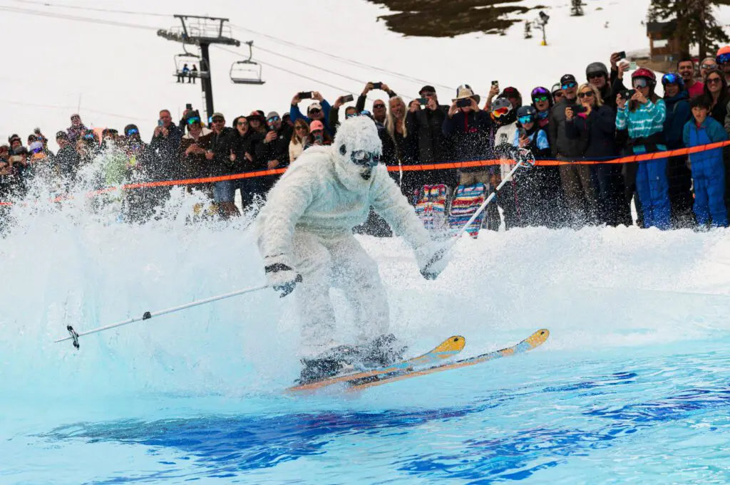 Mt. Rose A person dressed in a white Yeti costume skis across a pool of water surrounded by snow, creating a big splash. A large group of spectators, some taking photos, watches the scene. A ski lift and snowy hill are visible in the background.