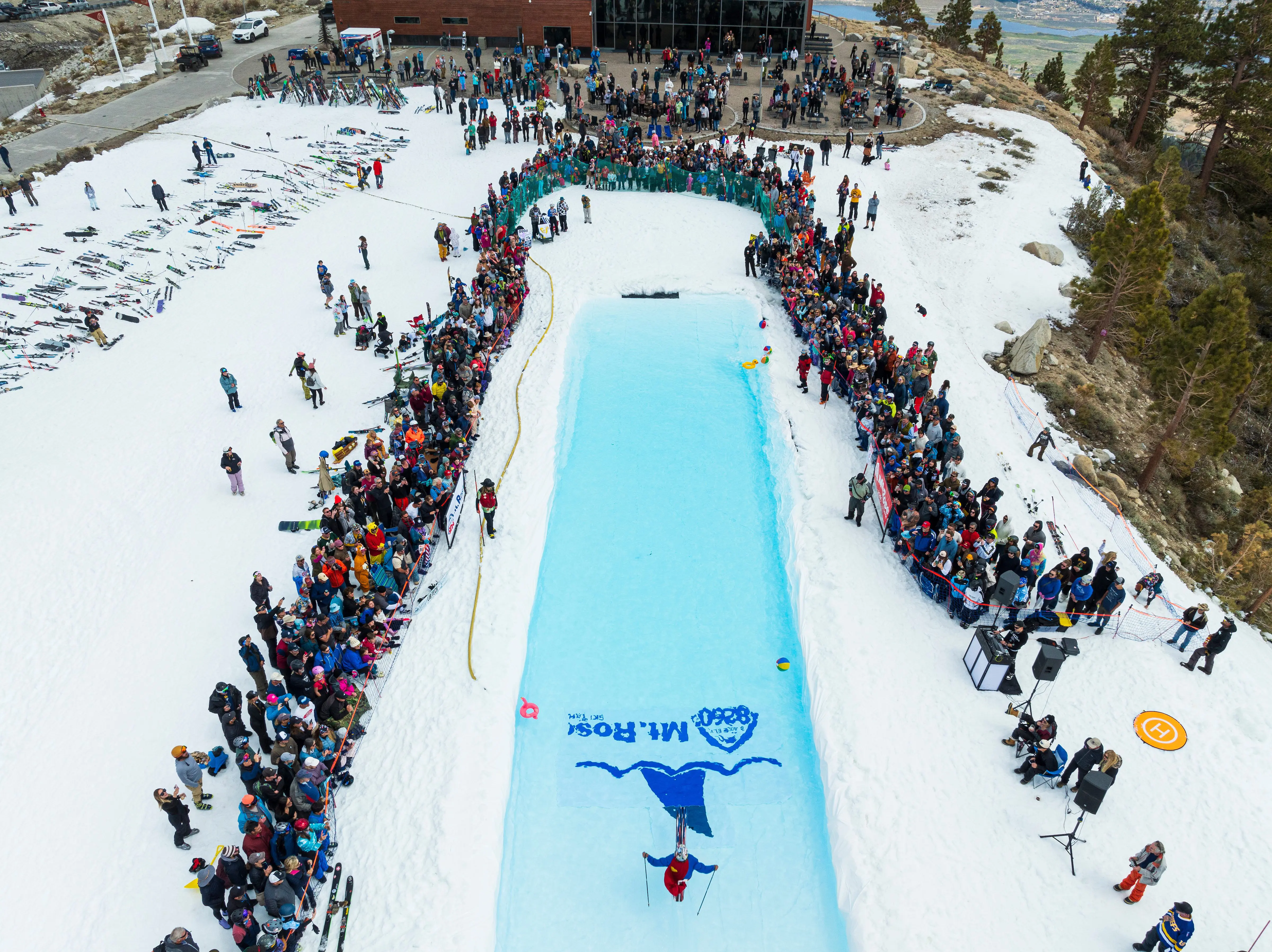 Mt. Rose An aerial view of a crowd gathered around a long, narrow pool set up in a snowy area. The pool is surrounded by people and skis. Some gathered people are dressed in ski gear while one person is skiing across the pool, surrounded by spectators on both sides.