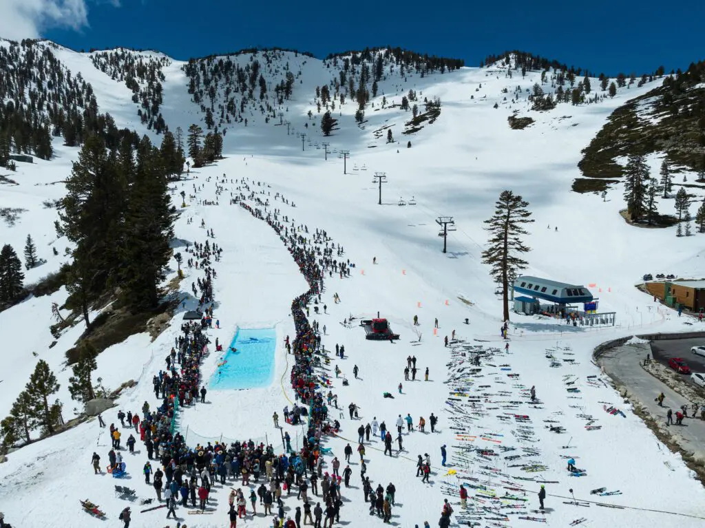 Mt. Rose A busy ski resort with a large crowd gathered around a rectangular pool on the snowy slopes of Slide Mountain. Skiers and snowboarders are in line for chairlifts, and ski equipment is laid out on the snow. Snow-covered trees and mountains form a picturesque backdrop under a blue sky near Winters Creek Lodge.