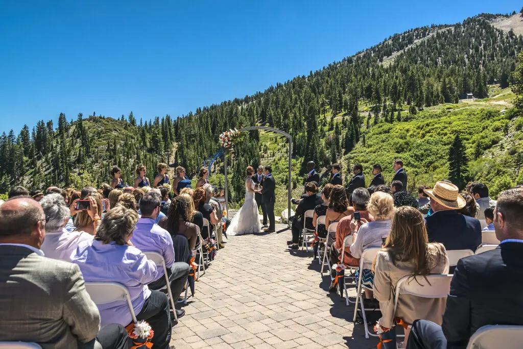 A mountain wedding ceremony unfolds under a clear blue sky, with guests seated on both sides of the aisle. The couple stands under a floral arch, framed by forested hills. Everyone is attentively watching this beautiful outdoor celebration. Mt. Rose Ski Tahoe