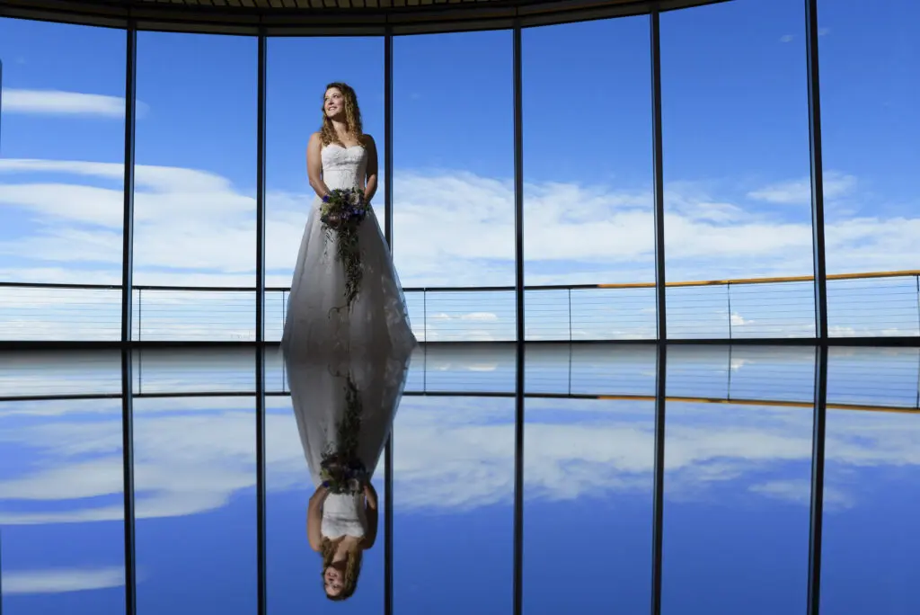 A bride in a white wedding gown stands smiling while holding a bouquet of flowers in a spacious room with large, curved windows showing a bright blue sky with clouds. Her reflection is clearly visible on the shiny floor below her. Mt. Rose Ski Tahoe