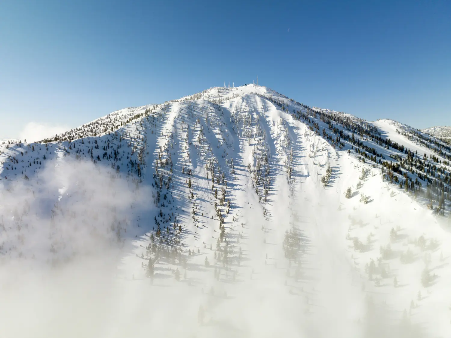A snow-covered mountain peak is bathed in sunlight against a clear blue sky. The mountain is dotted with sparse trees, and wisps of fog or low clouds drift near its base. Antenna structures are visible at the summit. Mt. Rose Ski Tahoe