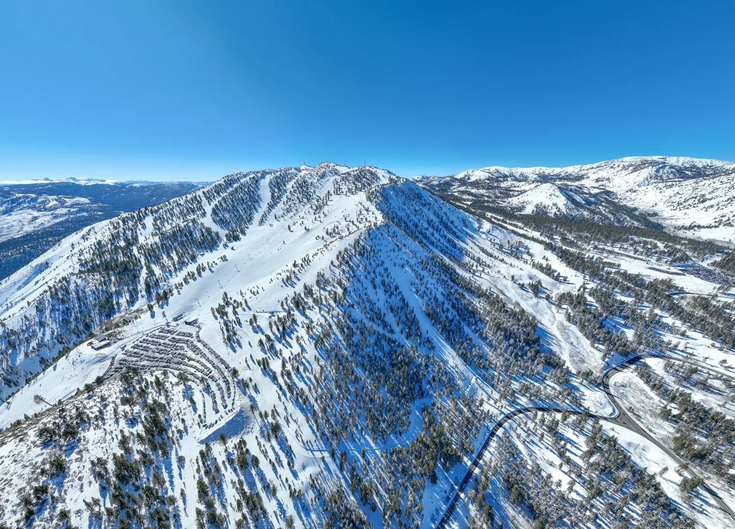 Aerial view of a snow-covered mountain range with ski slopes, surrounded by scattered trees. Clear blue sky above, winding roads and distant mountains visible in the background. Plenty of snow creates a winter wonderland scene. Mt. Rose Ski Tahoe