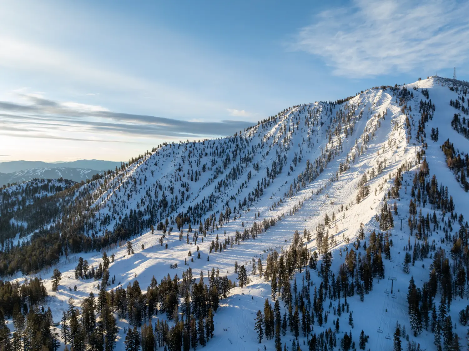 Aerial view of a snow-covered mountain with coniferous trees lining its slopes. The sun casts soft light, creating long shadows. The sky is mostly clear with a few scattered clouds, enhancing the serene, winter landscape. Ski runs and chair lifts are visible. Mt. Rose Ski Tahoe