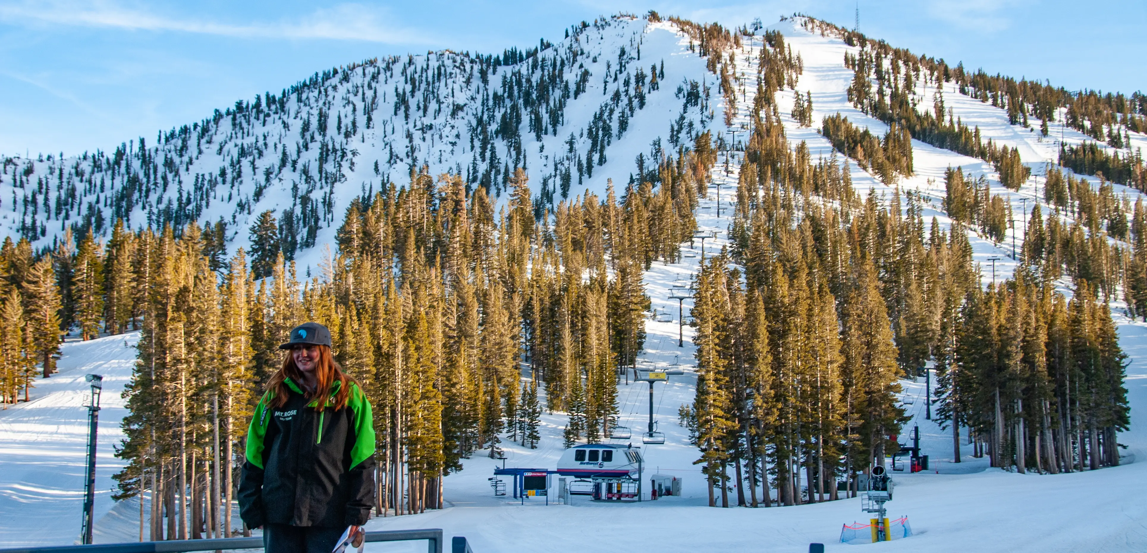 Woman in a green and black jacket and a blue cap stands on a snow-covered platform with a backdrop of a forest and a snow-laden mountain featuring ski trails. A ski lift is visible midway up the mountain, and the sky is clear. Mt. Rose Ski Tahoe