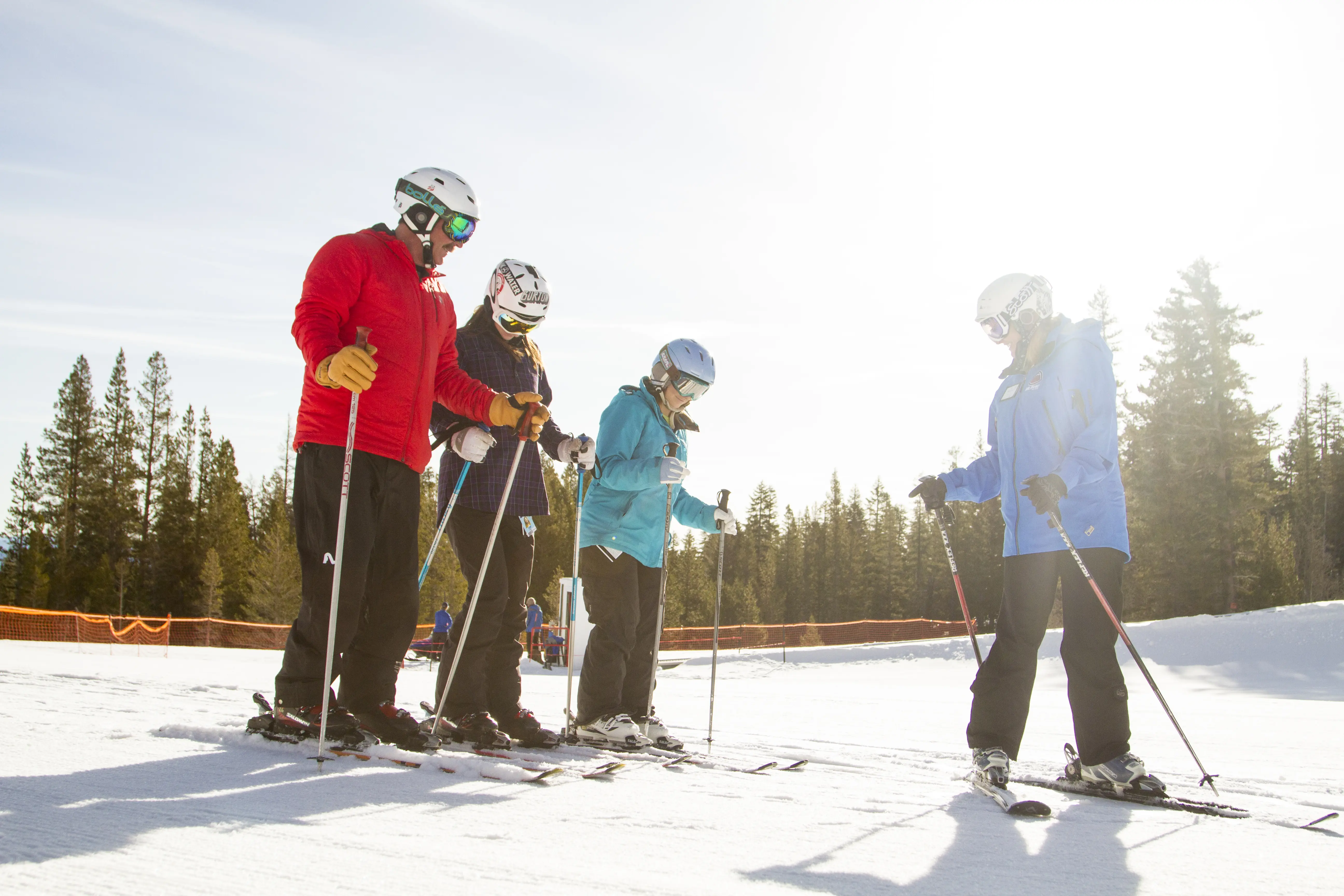 Four skiers dressed in winter gear stand on a snowy slope, adjusting their skis. They are wearing helmets and goggles. Tall pine trees and a clear blue sky are in the background, with sunlight casting a warm glow on the scene. Mt. Rose Ski Tahoe