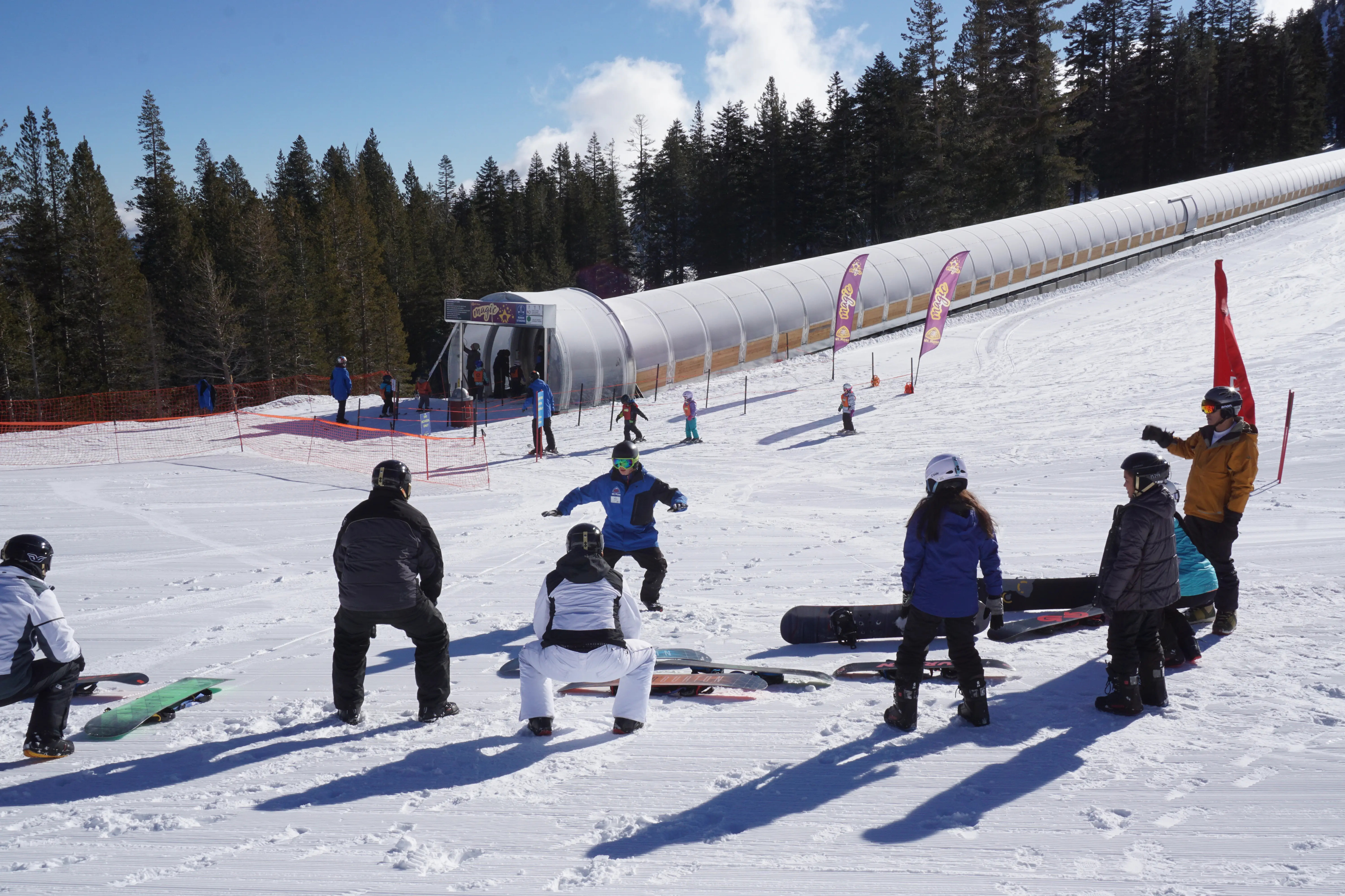 A group of people, some standing and some sitting on snowboards, gather on a snow-covered slope near a conveyor belt lift. Tall pine trees are in the background under a clear blue sky. A coach appears to be instructing the group, providing what could be the best kids ski lessons. Mt. Rose Ski Tahoe