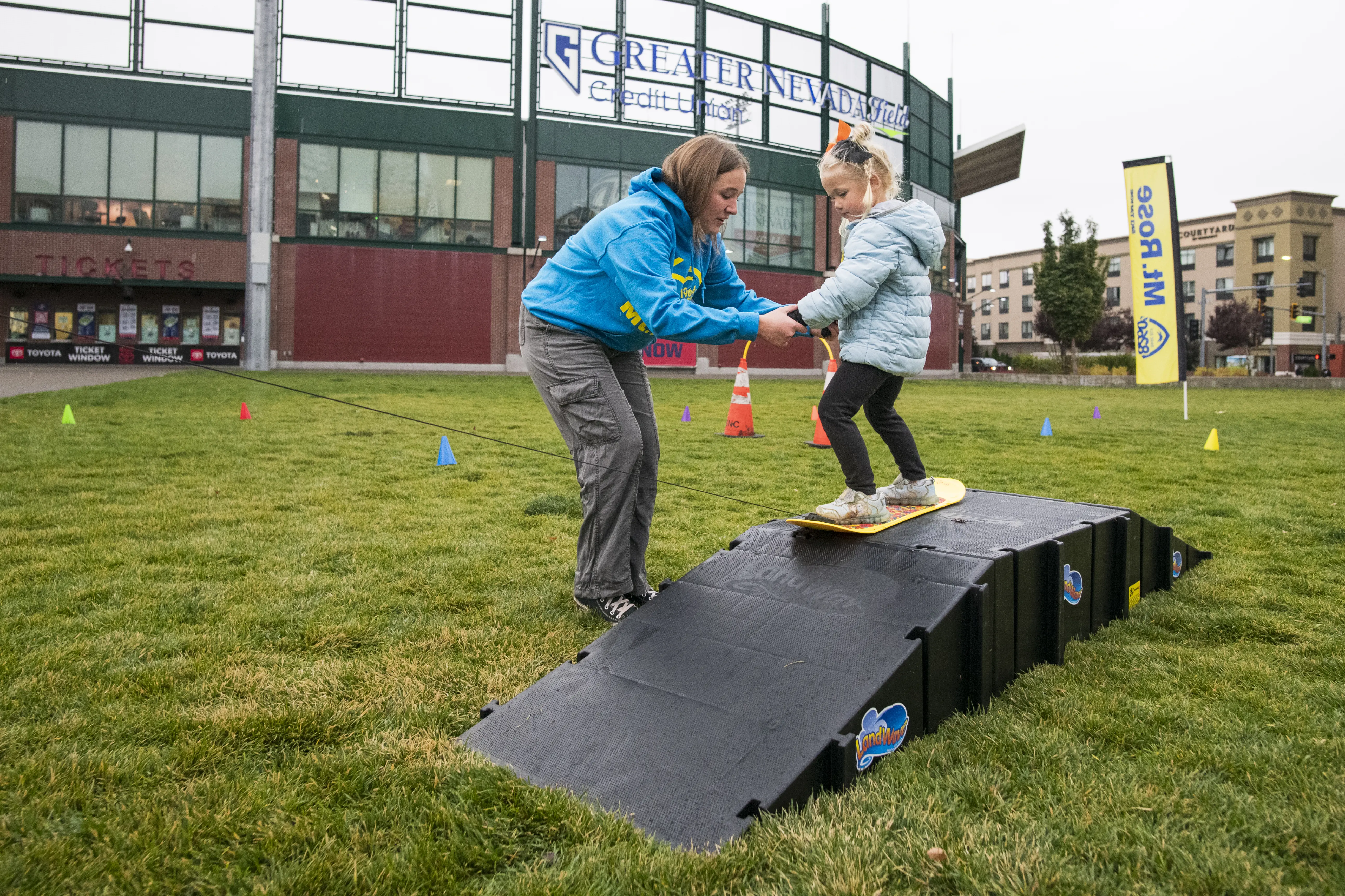 A woman helps a young girl balance on a small ramp with a snowboard on grass. They are outdoors near a building with banners and signs. Colorful cones are scattered around the area. Mt. Rose Ski Tahoe