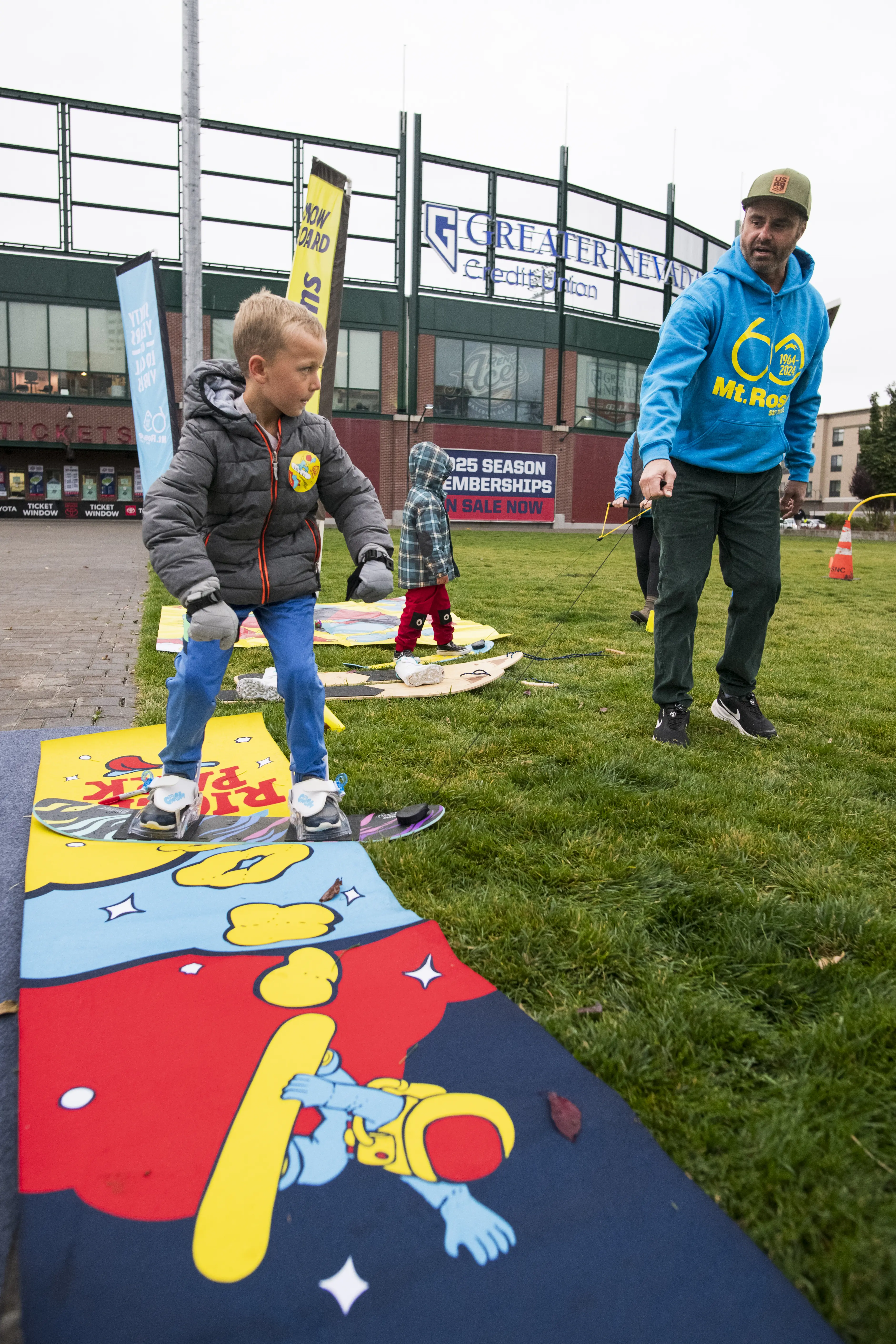 A young boy in a gray jacket practices snowboarding on a colorful mat with a cartoon design, guided by an adult in a blue hoodie. They are outdoors on grass, near a stadium or sports venue. Mt. Rose Ski Tahoe