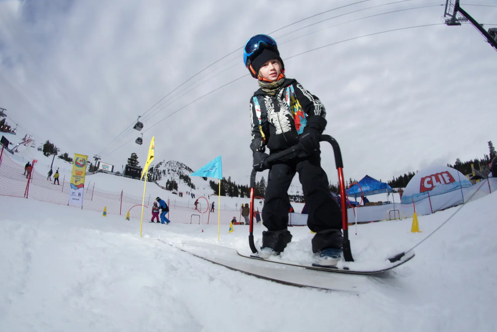 A child wearing a helmet and winter gear rides a snowboard on a snowy slope. There are colorful flags and ski lift cables in the background under a cloudy sky. Other people are also present in the skiing area. Mt. Rose Ski Tahoe