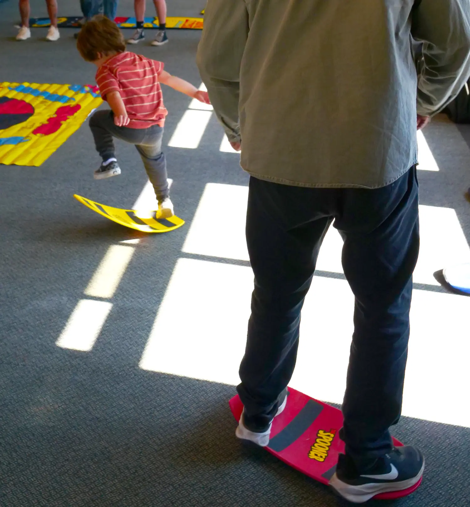 A child in a red striped shirt balances on a yellow board, while an adult in a green jacket and dark pants stands on a red and black board. They are indoors on a carpeted floor with sunlight streaming in. Mt. Rose Ski Tahoe