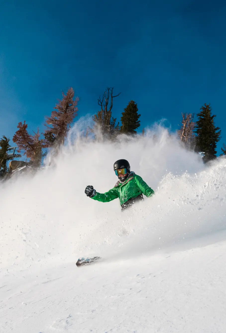 A skier in a light green jacket and black helmet moves swiftly downhill at Ski Tahoe, kicking up a spray of snow. They are surrounded by evergreen trees under a clear blue sky. Mt. Rose Ski Tahoe