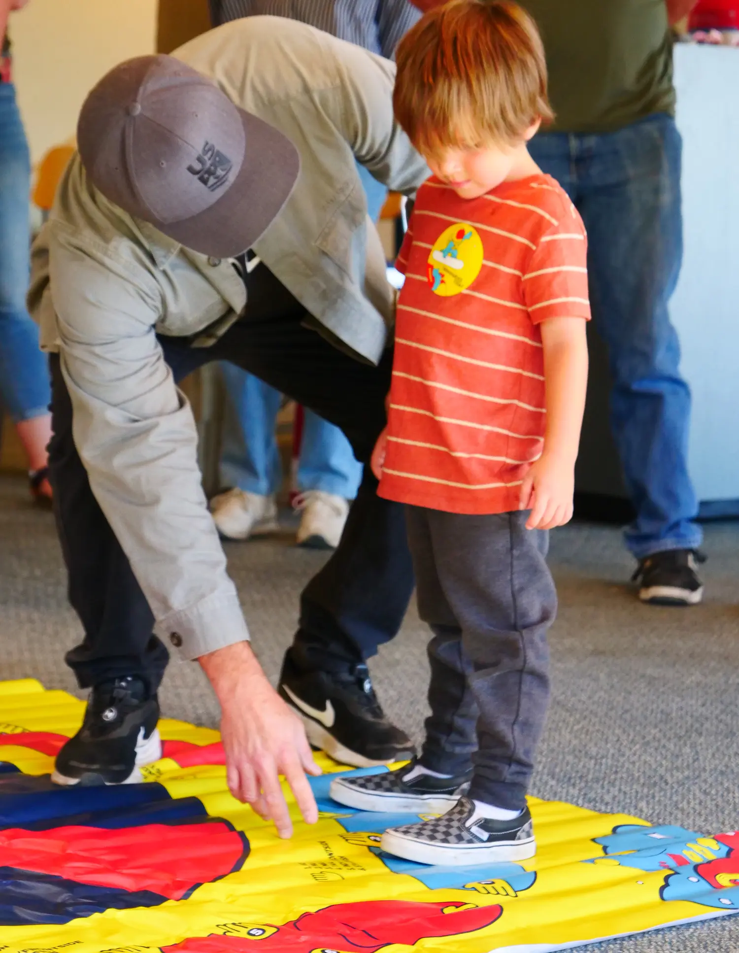 A child in a red striped shirt stands on a colorful map mat, while an adult in a grey jacket and cap kneels next to him, pointing at the map. Several people are in the background. Mt. Rose Ski Tahoe