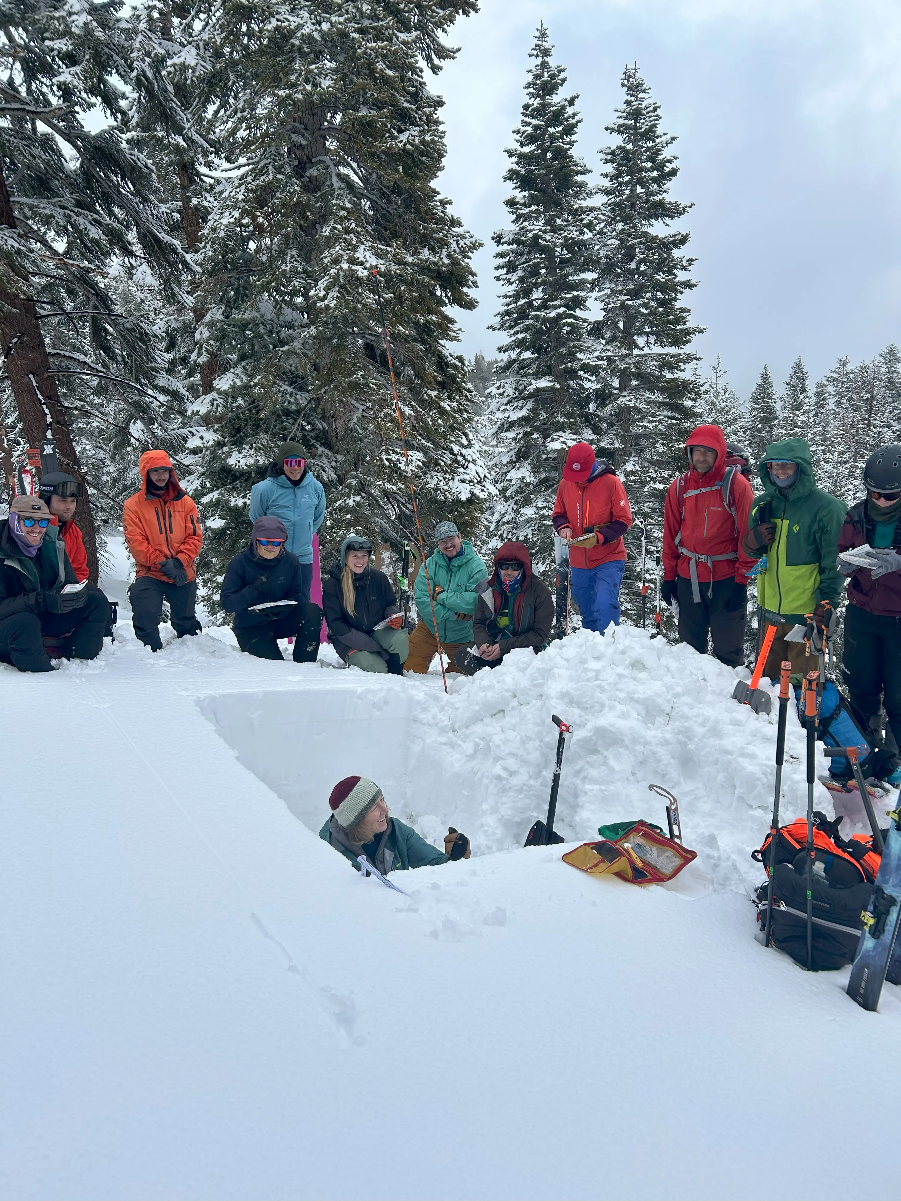 A group of people in winter clothing gather around a person sitting in a snow pit in a snowy forest. Various tools and gear are placed nearby. Trees covered in snow are in the background. Mt. Rose Ski Tahoe