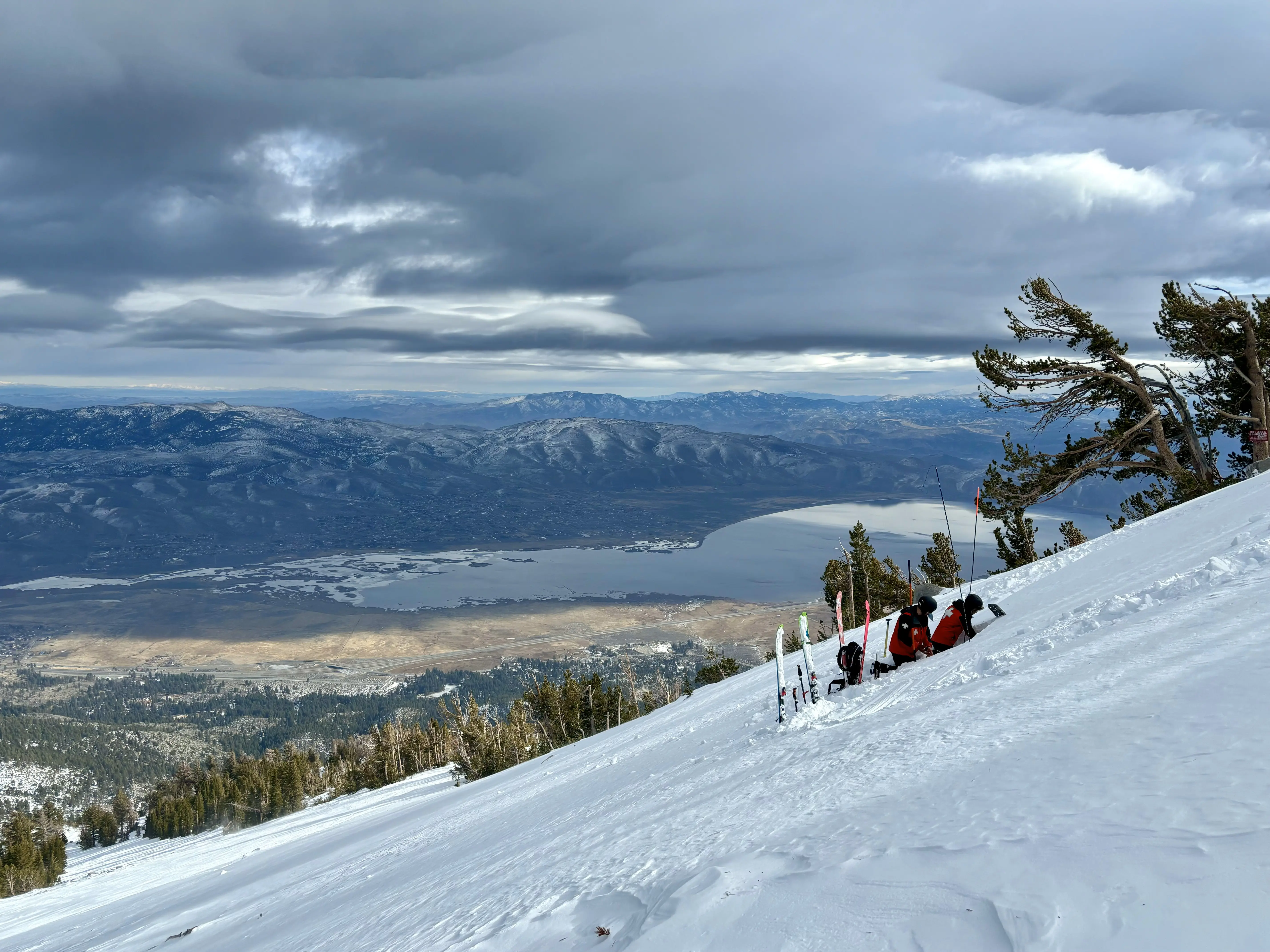 A snow-covered mountainside overlooks a vast landscape with a lake and distant mountains. Skis are propped up near a few resting skiers, and the sky above is filled with dramatic clouds. Mt. Rose Ski Tahoe